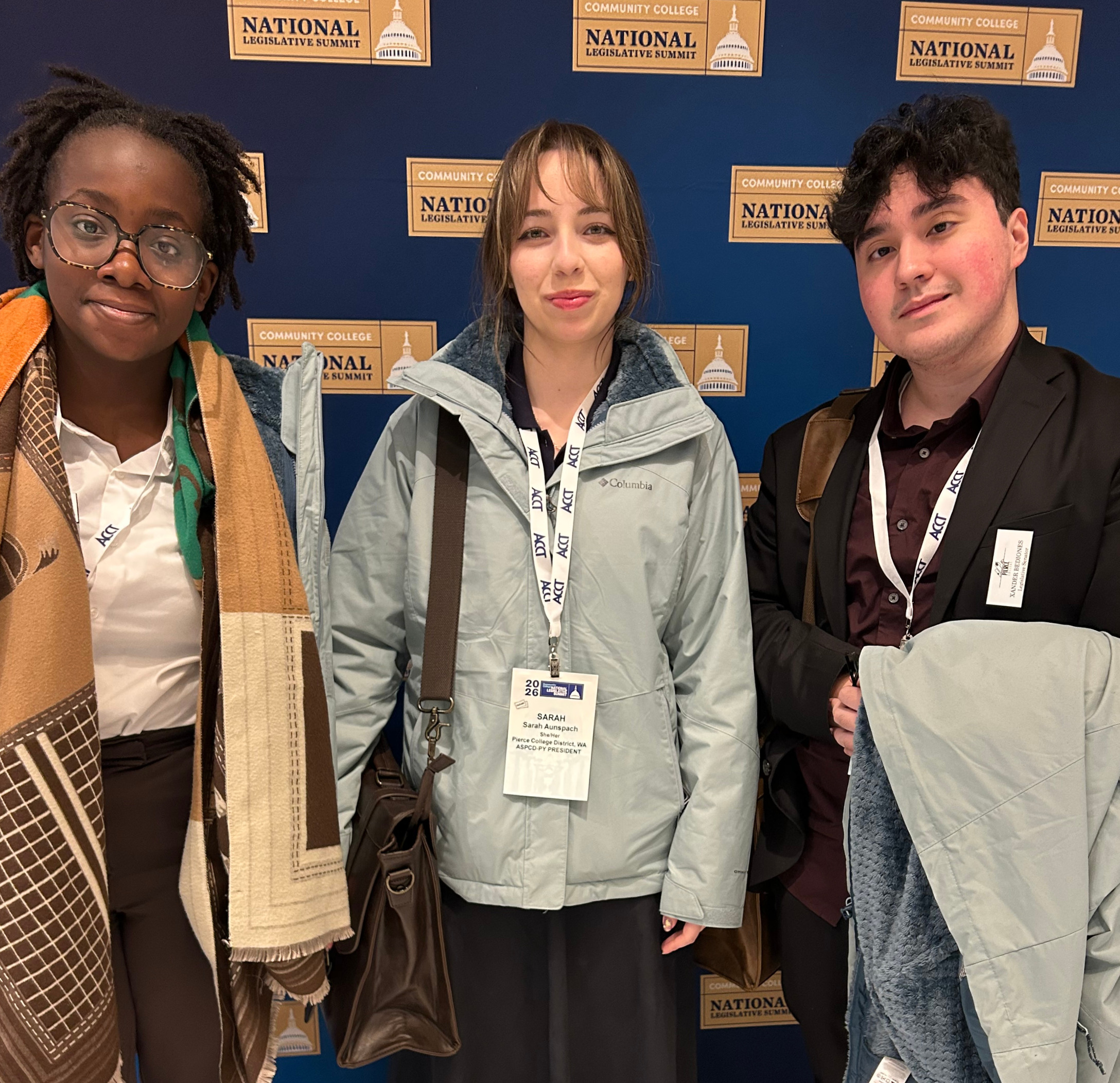 three students stand in front of a blue step and repeat with the association of community college trustees logo on it