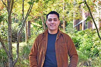 Vitaly Bravo Urrutia photographed in a brown jacket and blue shirt sitting in front of trees and the college center building