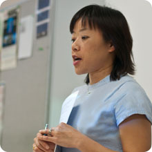 instructor speaking in front of classroom while holding a paper and pen